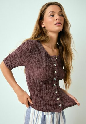 Sieviete ar gariem matiem, tērpusies teksturētā brūnā pogātas krekla un svītrainās biksēs uz vienkārša gaiša fona.