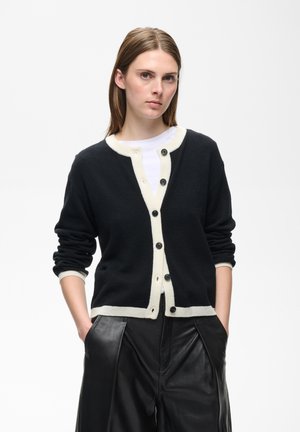 Jeune femme aux cheveux bruns et raides porte un cardigan noir avec une bordure blanche sur un haut blanc et un pantalon en cuir noir, les mains dans les poches.
