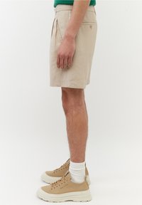 Shorts beige con tasche laterali, chiusura con bottone e taglio dritto; abbinati a sneakers color nocciola e calzini bianchi.