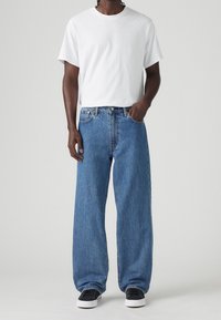 Man med enfärgad vit t-shirt, lösa blå jeans, svarta sneakers, står med ena handen i fickan mot en enfärgad bakgrund.