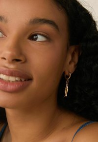 Øreringe formet som guldfisk med en hoops-design, der har en poleret guldfinish og en hængende fisk-accent. Hudtonen fremhæver smykkernes glans.