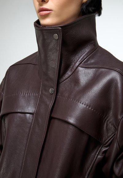 Közelkép egy személyről, aki sötétbarna bőrdzsekit visel, magas gallérral és patentos záródással.