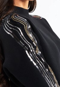 Maglione nero con strisce ondulate di paillettes argento, oro e nero sulla spalla e lungo le maniche, indossato da una persona con capelli lunghi.