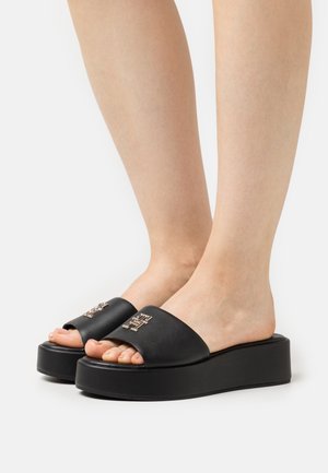 Feet wearing black platform slide sandals with metallic logo detail on the wide strap, against a white background.