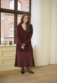 Maglione a maglia bordeaux con scollo a V, abbinato a una gonna in satin bordeaux. La modella indossa tacchi neri a punta. Fondo in legno e sfondo con finestra.