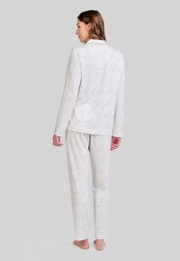 Mujer de pie descalza, vestida con pijama blanco con pequeños lunares azules y negros, de espaldas contra un fondo gris.