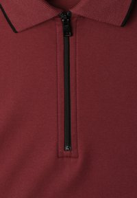 Gros plan d'une chemise en tissu bordeaux avec une fermeture éclair verticale noire et un col avec un bord noir.