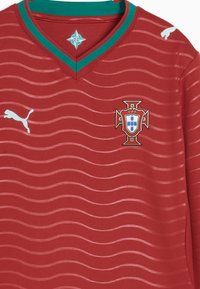 Camiseta roja de fútbol con patrón de ondas en rojo claro, cuello en V verde y rojo, logotipos de Puma y escudo de la selección nacional de Portugal en el pecho.