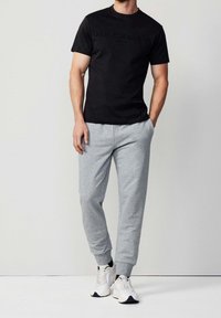 Czarna bawełniana koszulka z wytłaczanym logiem "HACKETT SPORT", zestawiona z szarymi spodniami dresowymi i białymi butami sportowymi z czarnymi akcentami.