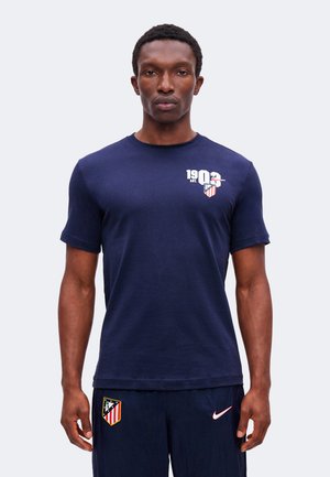 Homme portant un t-shirt bleu marine de l'Atlético Madrid et un short assorti avec le logo Nike, debout face à un fond uni.