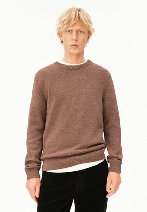 Blonder Mann trägt einen braunen Strickpullover über einem weißen Hemd und steht vor einem schlichten weißen Hintergrund.