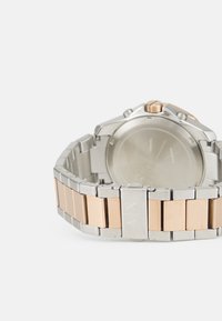 Silber- und roségoldfarbene Edelstahl-Uhr mit einem Gliederarmband, rundem Gehäuse, Krone an der Seite und Logo am Verschluss. Minimalistisches Design.