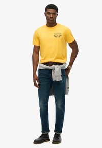 Camiseta de algodón amarilla con un estampado gráfico, combinada con unos jeans de mezclilla azul y un suéter gris claro atado a la cintura. Zapatos negros.