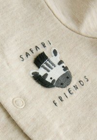 Izvezena glava zebre s tekstom "Safari prijatelji" na bež tkanini, pored kopče na dječjem odjevnom predmetu.