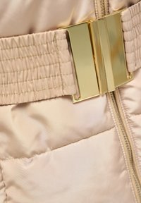 Tejido acolchado beige con un acabado brillante, que presenta un amplio detalle de fruncido y un cierre de hebilla de metal dorado. Cremallera en el centro.