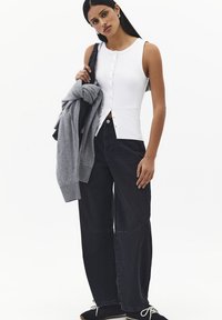 Mujer con cabello largo y oscuro que lleva una blusa blanca sin mangas con botones, vaqueros negros de pierna ancha y sostiene un jersey gris sobre un hombro.