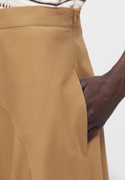 Χέρι που ακουμπά στην πλαϊνή τσέπη μιας ψηλόμεσης φούστας σε καμηλό καφέ χρώμα με φερμουάρ στη μέση, φορεμένη με κρεμ πλεκτό τοπ.