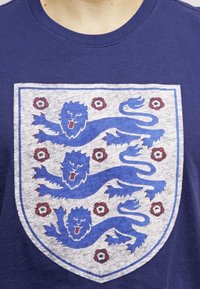 Marineblaues T-Shirt mit einem weißen Schild, das drei blaue Löwen und rote florale Akzente zeigt. Kurze Ärmel, runder Hals und Baumwolltextur.