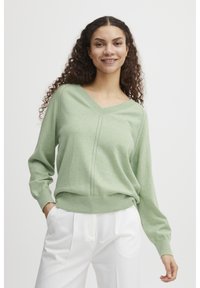 Hellgrüner V-Ausschnitt-Pullover mit gerippten Bündchen und Saum, der gestrickte Details entlang der Mitte aufweist. Kombiniert mit weißen Hosen.