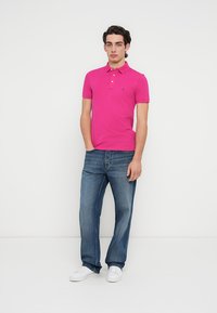 Ροζ πόλο μπλούζα με λαιμόκοψη με τρία κουμπιά, συνδυασμένη με τζιν παντελόνι με μέτρια πλύση και λευκά αθλητικά παπούτσια. Απλή, καθημερινή ενδυμασία.