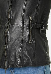 Schwarze Lederjacke mit strukturierter Oberfläche, aufwendiger Nahtführung, zwei seitlichen Reißverschlüssen und einem Schnallen-Detail an der Taille.