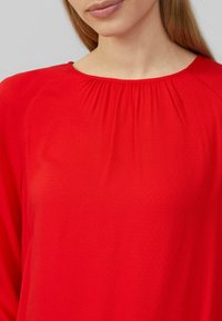 Blouse rouge présentant une texture douce et lisse, avec un col rond et des plis subtils aux épaules. Manches longues et tissu léger.