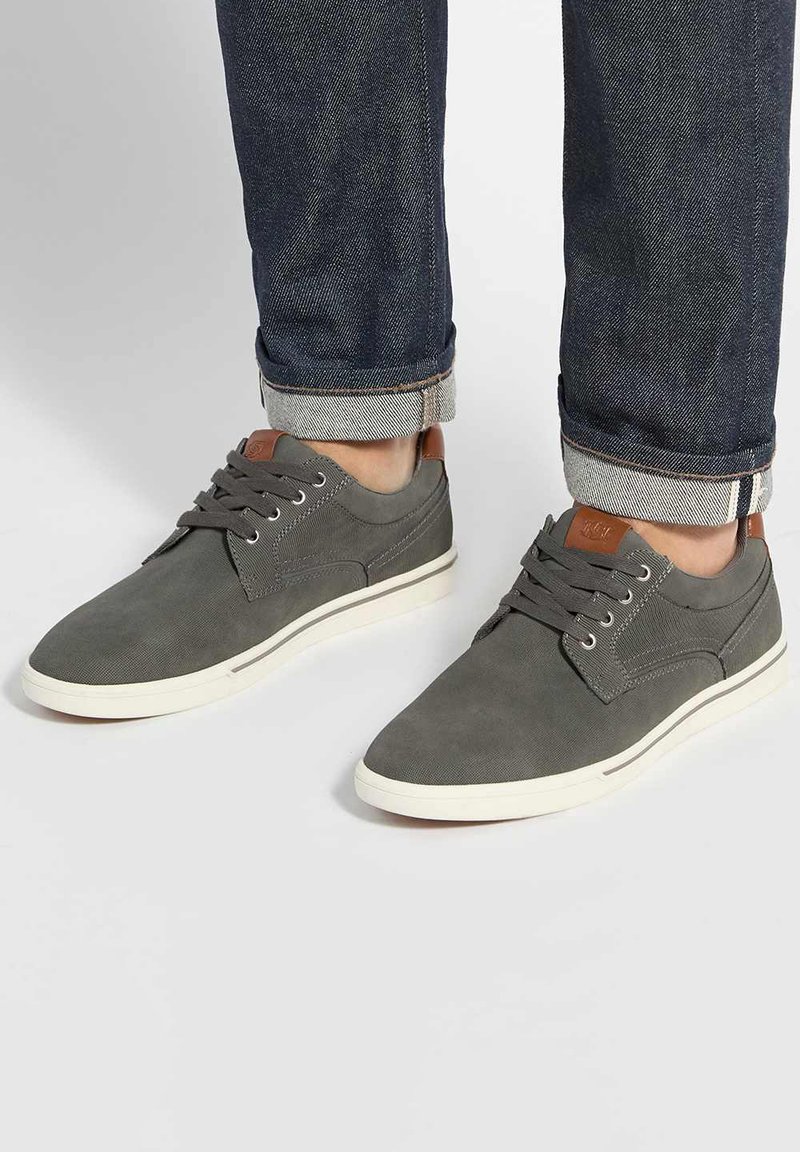 Chaussures décontractées grises à lacets portées avec un jean en denim foncé retroussé sur un fond clair et uni.