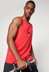 Rdeča majica brez rokavov iz bombaža s krožnim izrezom, na prsih pa ima majhen črn logotip Adidas. Model drži utež.