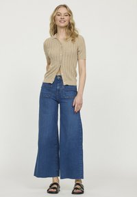 Cardigan beige à manches courtes avec texture côtelée, devant à boutons, associé à un jean bleu à jambes larges et des sandales noires, peu de détails.