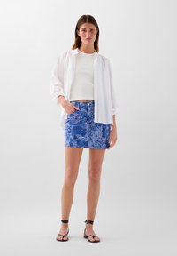 Camicia bianca oversize, canotta bianca a coste, mini gonna in denim blu con stampa, e sandali neri con cinturini, il tutto su uno sfondo semplice.
