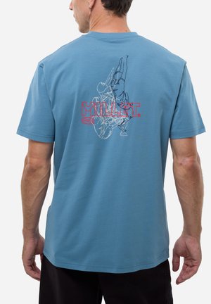 Homme portant un t-shirt bleu avec un dessin en ligne d'un grimpeur s'étirant vers le haut et un logo rouge de la marque "MILLET" au dos.