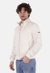 Giacca bomber leggera di colore crema con colletto alto, chiusura con zip, tasche laterali e polsini a coste. Presenta il logo sulla manica.