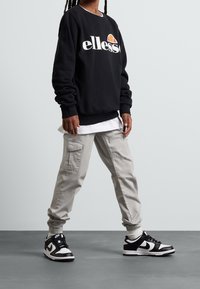 Felpa nera con logo bianco, indossata sopra una camicia bianca. Pantaloni cargo grigi chiari e sneaker bianco e nero completano l'outfit.