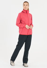 Lätt rosa jacka med dragkedja fram och justerbar huva, kombinerad med mörka byxor. Jackan har fickor med dragkedja och elastiska cuffs.