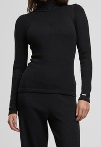 Zwarte langarm pullover met hoge kraag, geribbelde textuur en een halve rits. Heeft een subtiele logotag bij de manchetten.