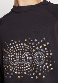 Sudadera negra con cuello redondo, que presenta un diseño de "HUGO" con tachuelas en dorado, plateado y rojo en la parte frontal, con un cuello acanalado texturizado.