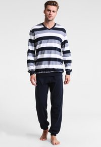 Man staat blootsvoets in een gestreepte langslokige shirt en donkere joggingbroek tegen een eenvoudige lichte achtergrond.