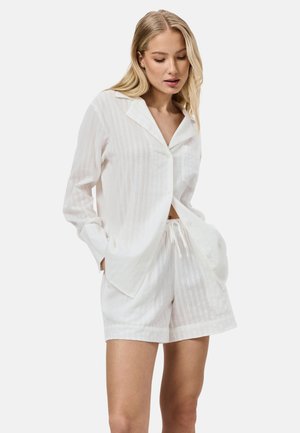 Blondine vrouw draagt een wit lange mouwen overhemd met kraag en bijpassende short met trekkoord, handen in de zakken, kijkt naar beneden.