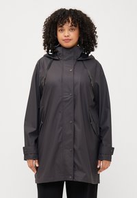 Woman with curly hair wearing a dark waterproof jacket with a hood, standing against a plain white background.