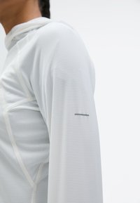 Κοντινό πλάνο ενός ατόμου που φοράει λευκό πουκάμισο μακρυμάνικο με υφή, εμφανείς γραμμές ραφής και μια μικρή οριζόντια μαύρη ρίγα στο μανίκι.