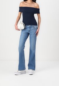 Top azzurro scuro con spalle scoperte, jeans a zampa d'elefante azzurro chiaro, sneakers bianche e borsa bianca. La texture del denim e il busto aderente mettono in risalto la silhouette.