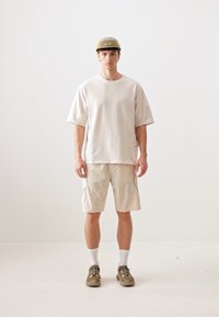Beige oversized t-shirt with short sleeves, paired with knee-length beige shorts. Worn with a light green cap and gray hiking shoes.