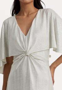 Kvinna klädd i en klänning av silvermetallisk tyg med djup V-ringning och vriden knutdetalj i midjan, med breda svängande ärmar.