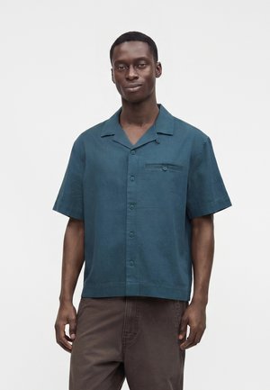 Homme portant une chemise à manches courtes coloris vert sarcelle foncé et un pantalon marron, debout devant un fond clair uni.