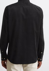 Camisa de pana negra con cuello punta, mangas largas y puños abotonados. Presenta una textura suave y un dobladillo curvado.