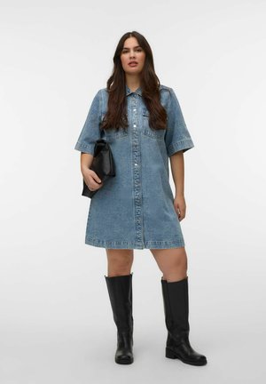 Ung kvinde med langt hår iført en knappet kortærmet denimkjole, der holder en sort clutch og bærer knælange sorte støvler.