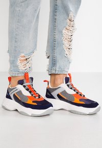 Nohy v mnohobarevných oranžových, námořnických, šedých a bílých teniskách doplněných o světle modré roztrhané džíny na čistém bílém pozadí.