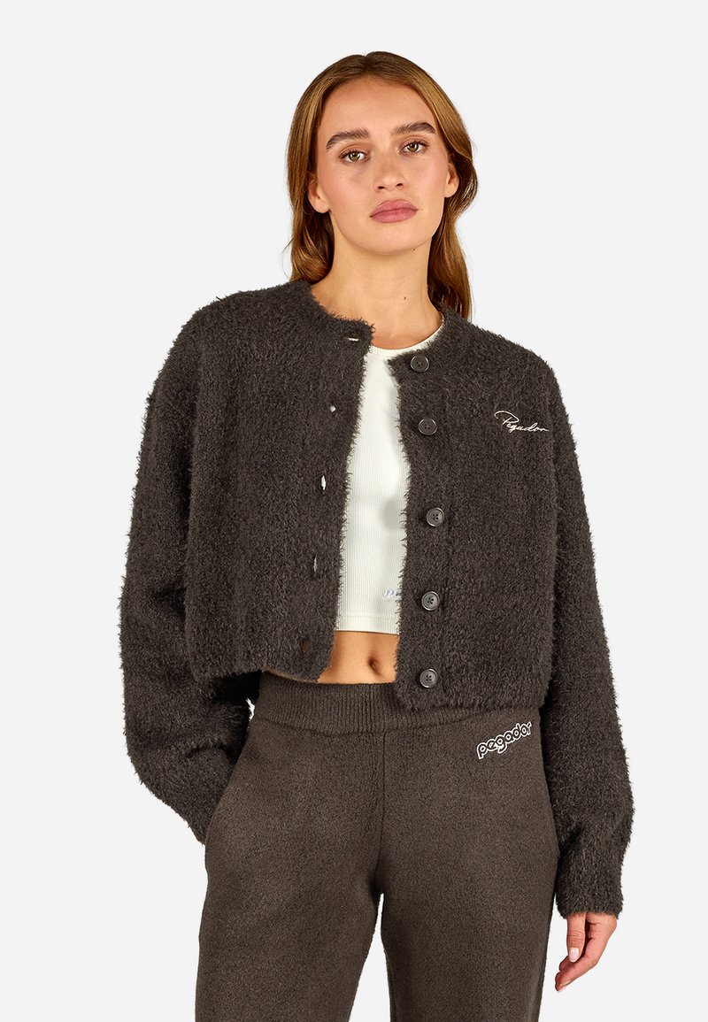 Šedý, zkrácený kardigan z měkké texturované látky. Má velké černé knoflíky a kulatý výstřih. Modelka ho nosí přes bílé tričko.