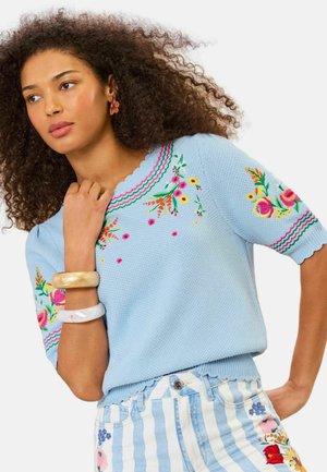 Kvinde med krøllet hår iført en lyseblå sweater med blomsterbroderi og stribede bukser med blomsterlapper, accessoriseret med armbånd.