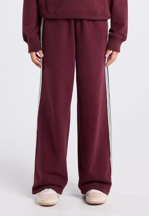 Bordeauxrote Sweatpants mit weitem Bein und weißen Seitenstreifen, kombiniert mit passendem Sweatshirt und beigen Sneakers auf hellem Hintergrund.